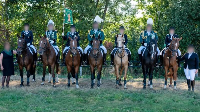 Sieben Reiter in Schützenuniform auf braunen, schwarzen und hellen Pferden stehen in einer Reihe vor Bäumen, in der Mitte eine grüne Vereinsfahne.